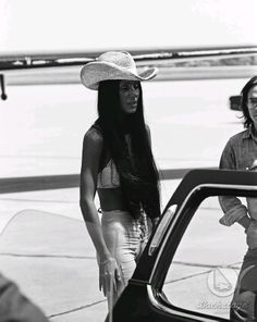 a woman in a cowboy hat standing next to a car and another man looking at her