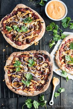 two pizzas with different toppings sitting on top of a wooden table next to a bowl of dipping sauce