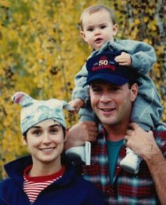 a man holding a baby wearing a hat while standing next to a woman in front of trees