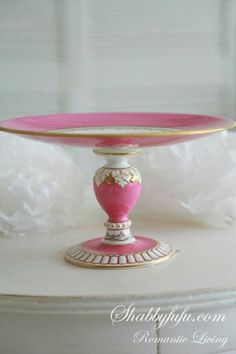 a pink and gold plate sitting on top of a white cake stand with flowers in the background