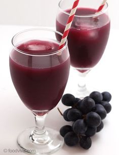 two glasses filled with red wine next to some grapes and a striped straw on a white table