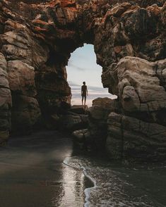 a person standing on the beach looking out at the ocean through an arch in the rocks