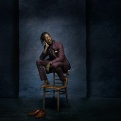 a man sitting on top of a wooden chair in front of a blue wall and wearing a purple suit