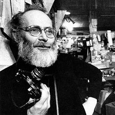 a man with a beard and glasses holding a camera in his hand while standing next to a wall full of books