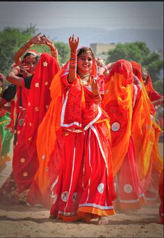 a group of women dressed in bright colored clothing dancing on dirt ground with trees in the background