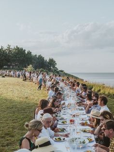 a group of people sitting at a long table with food on it in front of the ocean