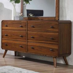 a wooden dresser sitting next to a white vase with flowers in it and a mirror on the wall