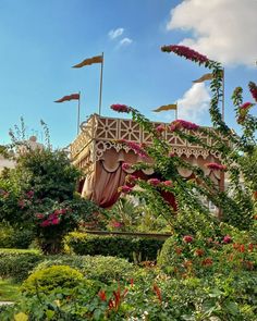 a garden area with flowers, bushes and a wooden bridge in the middle that has flags flying above it