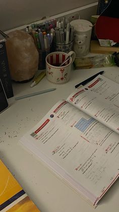 an open book sitting on top of a white desk next to a cup and pen