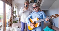 two people standing in a living room with a guitar and cell phone to their ear