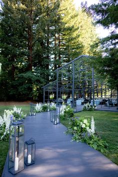 an outdoor walkway with lanterns and flowers on the side, surrounded by tall pine trees