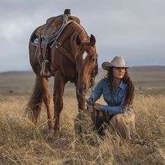 a woman kneeling down next to a brown horse on a dry grass field with another horse in the background