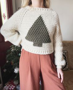 a woman standing in front of a christmas tree wearing a white sweater and brown pants