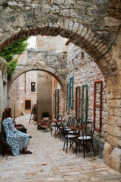 a woman sitting on a bench in an alleyway with tables and chairs around her