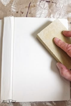 a person holding a piece of wood in their hand over an open book that is covered with white paper