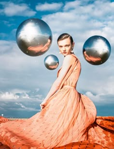 a woman in an orange dress is sitting on a rock with two silver balls floating above her