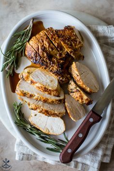 a white plate topped with sliced chicken next to a knife and some green herbs on top of it