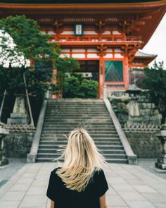 a woman with blonde hair standing in front of a red and orange building, looking up at the stairs