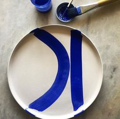 a white plate with blue paint on it next to two brushes and one empty cup