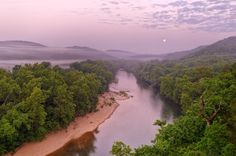 an aerial view of a river surrounded by trees and mountains in the distance at dusk