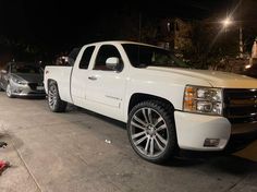 a white truck is parked on the side of the road at night with other cars behind it