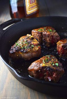 steaks cooking in a skillet on a wooden table next to a bottle of beer