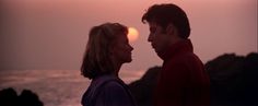 a man and woman standing next to each other near the ocean at sunset or sunrise