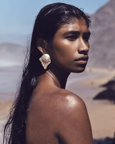 a woman with long hair and earrings standing in front of a mountain range, looking off into the distance