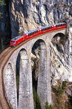 a red train traveling over a bridge on the side of a rocky mountain range in europe