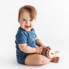 a smiling baby sitting on the floor with a cup in his hand and wearing a blue shirt