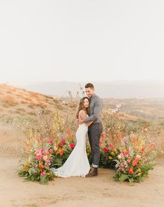 a bride and groom standing in front of flowers on the side of a hill at sunset