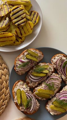 two plates with sandwiches and pickles on them next to some potato wedges in the background