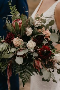 a bride holding a bouquet of flowers and greenery with her groom in the background