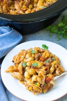 a white plate topped with pasta next to a slow cooker filled with meat and veggies