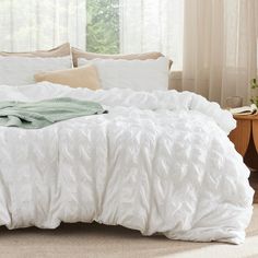 a bed with white comforter and pillows in a room next to a window,