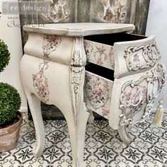 an ornately decorated white cabinet with drawers and knobs on the doors is next to a potted plant