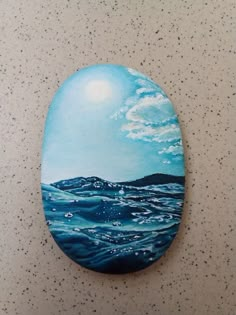 an oval painted rock with water and clouds on it, sitting on a countertop