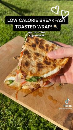 a person holding a sandwich on top of a wooden cutting board in the middle of grass