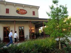 two people walking in front of a restaurant with trees and bushes around it on a sunny day