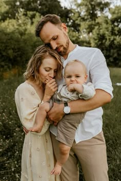 a man and woman holding a baby in their arms while standing in the grass with trees behind them