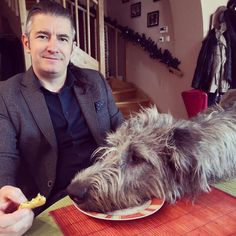 a man sitting at a table with a dog eating food from a plate on his lap