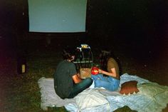 two people sitting on a blanket in front of a projection screen at night with an apple