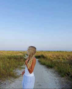 a woman walking down a dirt road next to tall grass