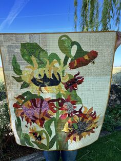 a woman holding up a quilt with flowers on it in front of some grass and trees