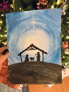 a hand holding up a christmas card with a nativity scene on it and a tree in the background