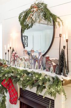 a mantel decorated with christmas greenery, candles and houses on top of it