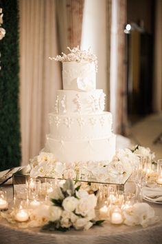 a white wedding cake sitting on top of a table covered in flowers and candles next to wine glasses