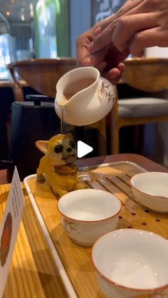 a person pouring tea into white cups on top of a wooden table with plates and silverware