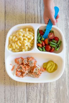 a child's hand holding a blue plastic spoon over a white plate filled with food