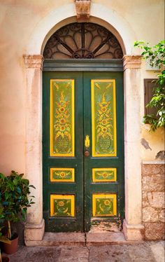 an ornate green door with yellow panels on the front and side doors is flanked by potted plants